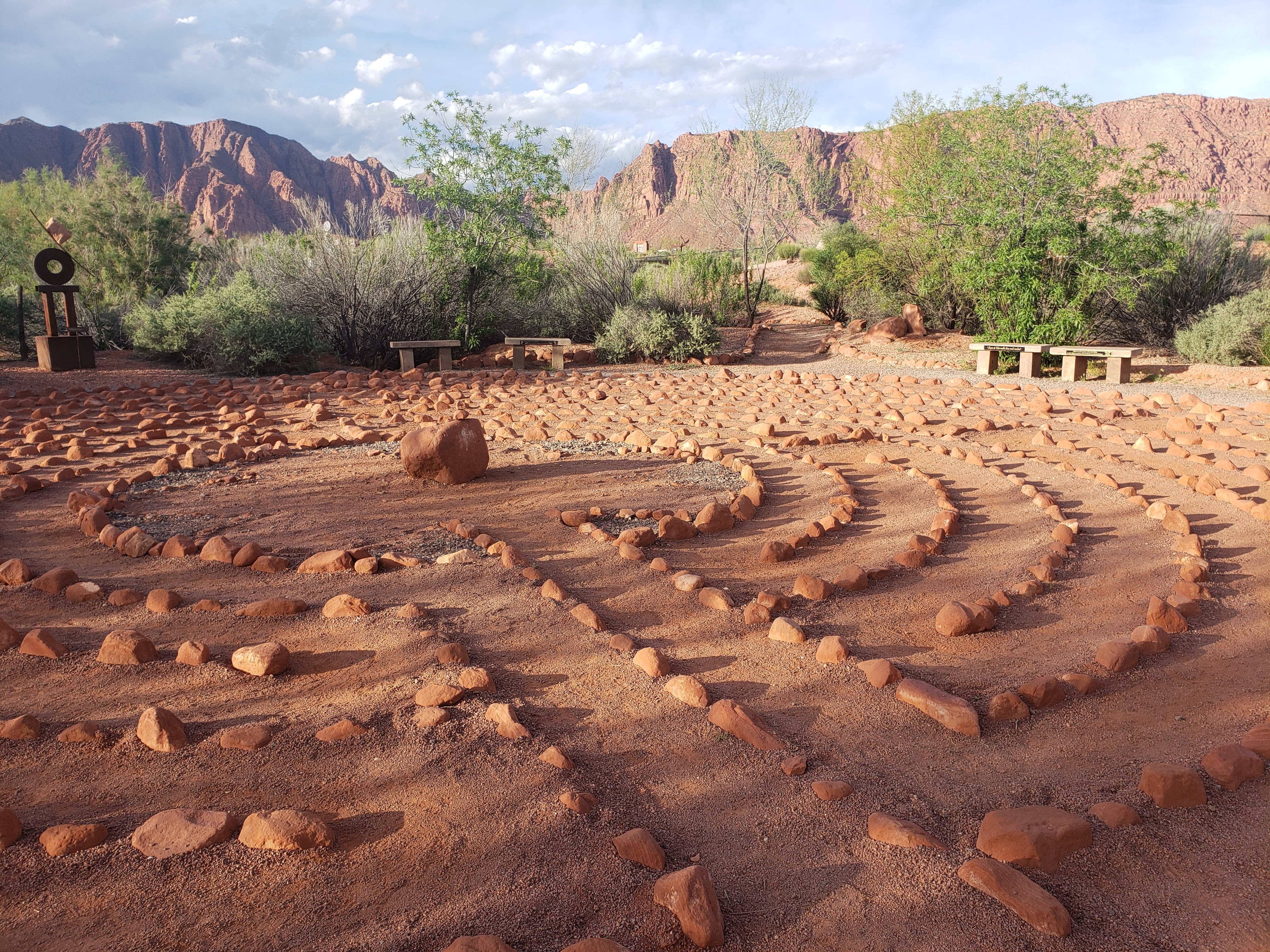 A stone labyrinth laid into red desert sand, surrounded by sage and ochre brush with mountains in the distance.