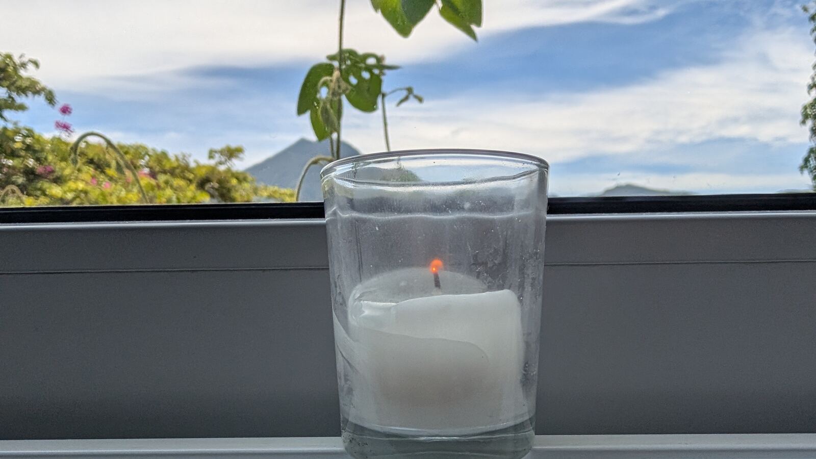 A candle in a glass on a windowsill beside a young plant, the lake and mountains beyond