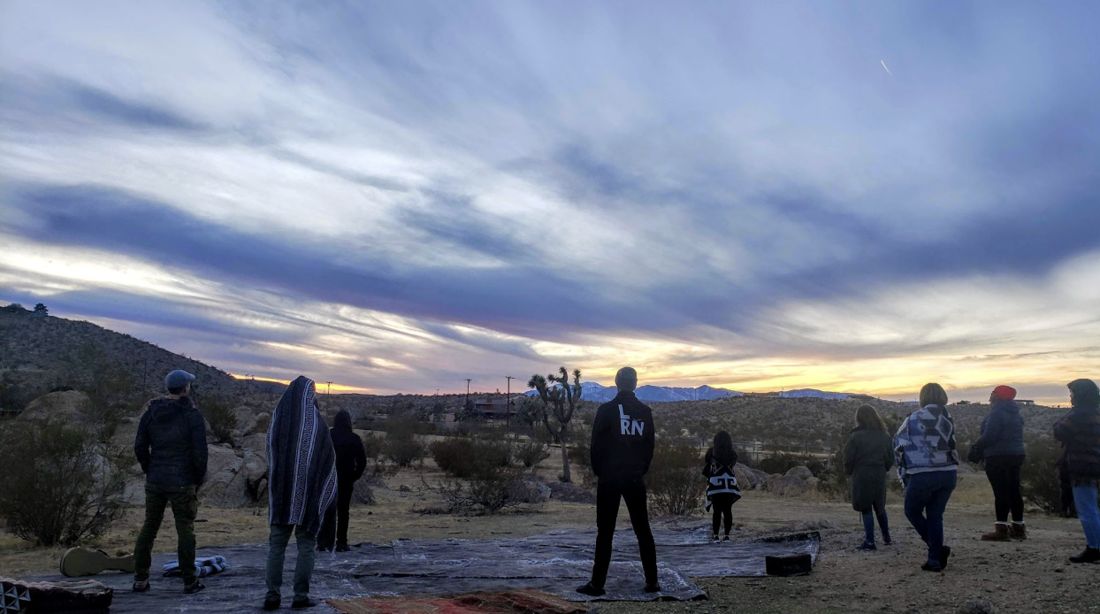 A small group standing on a desert hillside at dawn, facing a wide sky of streaked clouds.